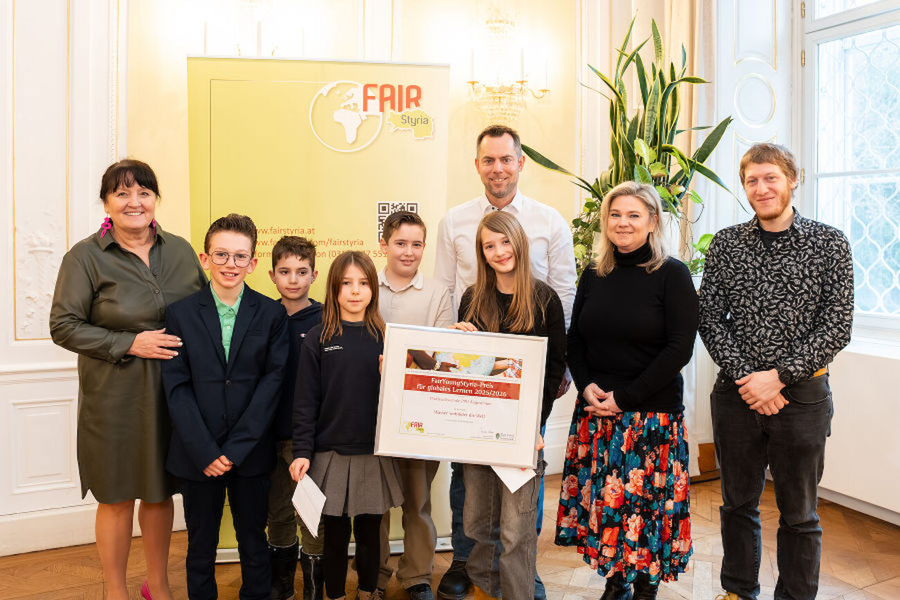 Preisverleihungsfoto, wo fünf Kinder, zwei Lehrer sowie die Schulleitung mit Manuela Khom in der Grazer Burg mit der erhaltenen Urkunde posieren.