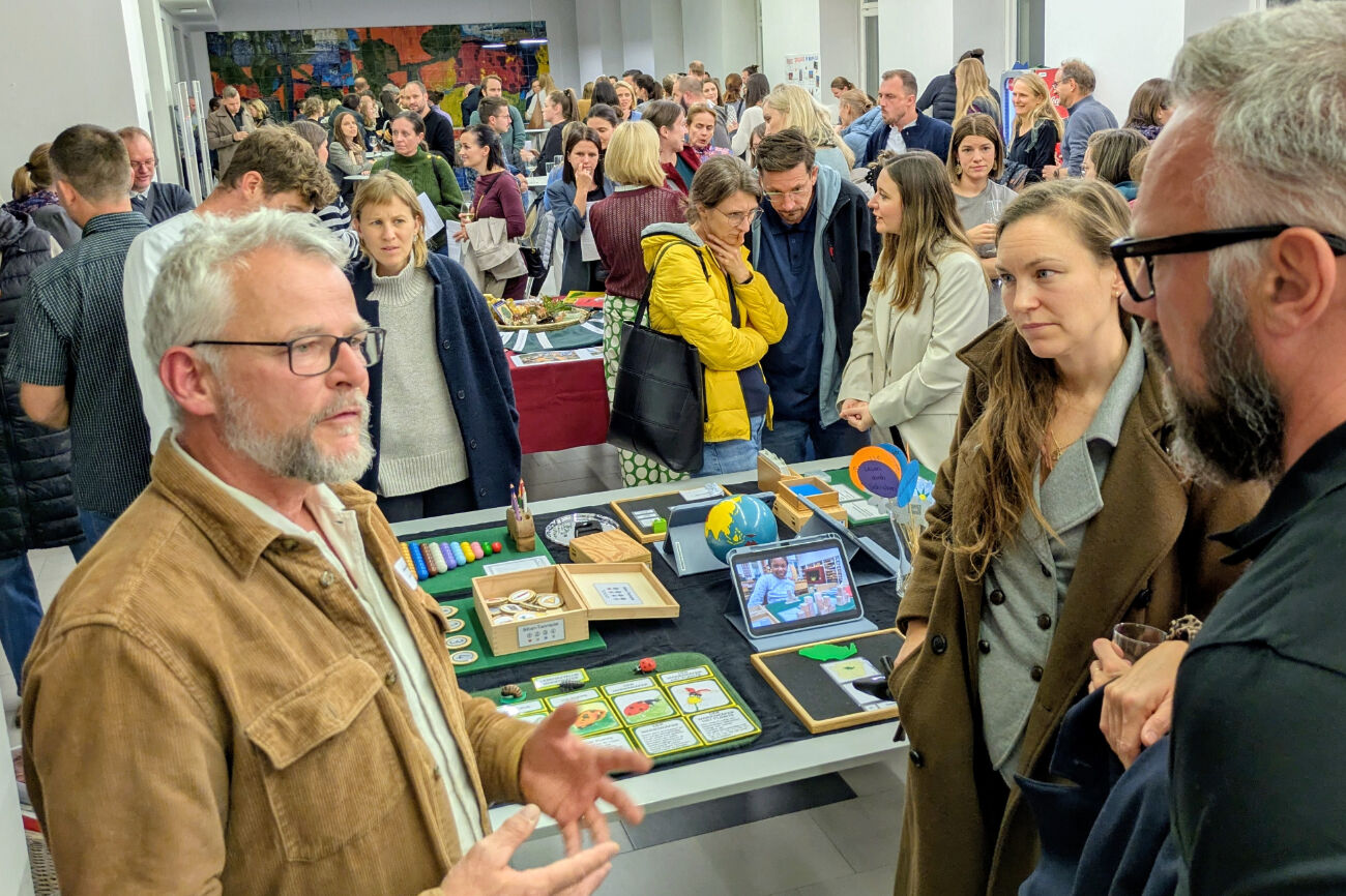 Kinder sitzen um einen Tisch herum und hören zwei Erwachsenen zu, die den Umgang mit Kräutern demonstrieren.