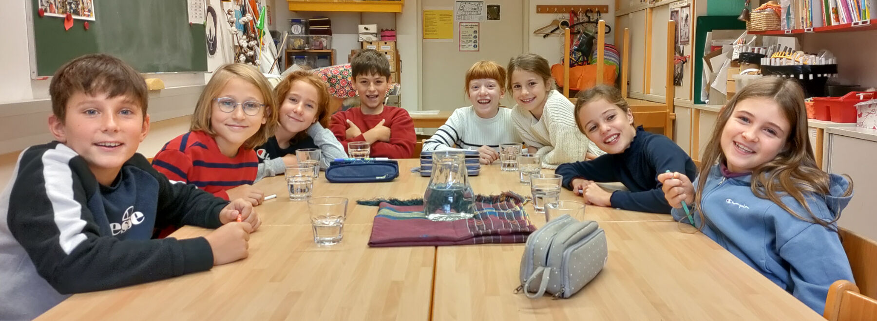 Acht Kinder sitzen an Holztischen bei der Schulratsitzung. Vor ihnen befinden sich Wassergläser.