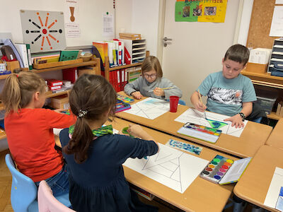 Vier kleine Kinder sitzen an Schreibtischen und malen geometrische Muster auf Papier.