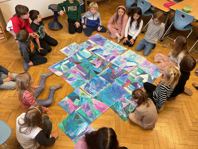 Kinder sitzen im Kreis um bunte abstrakte Kunstwerke auf dem Boden eines Klassenzimmers.