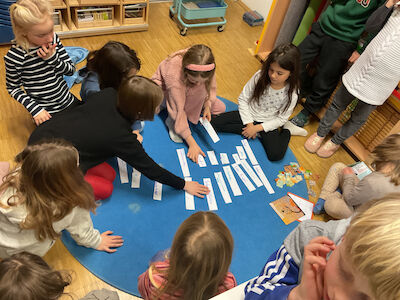 Kinder sitzen im Kreis auf einem blauen Teppich und ordnen Papierstreifen aneinander.