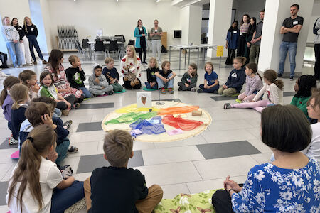 Kinder sitzen in einem hellen, offenen Raum im Kreis um ein buntes Tuch.