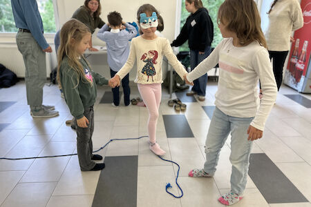 Drei Kinder halten sich an den Händen und bilden einen Kreis, eines trägt eine blaue Maske und steht auf einem Seil.