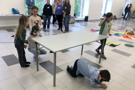 Vier Kinder spielen drinnen um einen Tisch herum, während Erwachsene im Hintergrund zuschauen.