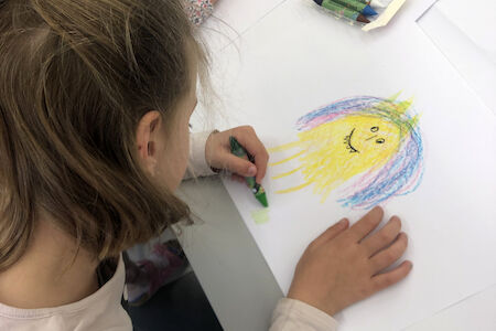 Kind zeichnet mit Buntstiften ein buntes lächelndes Gesicht auf weißes Papier.