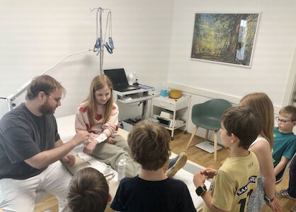 Ein Arzt und ein junges Mädchen unterhalten sich, während mehrere Kinder in einer Arztpraxis zuschauen.