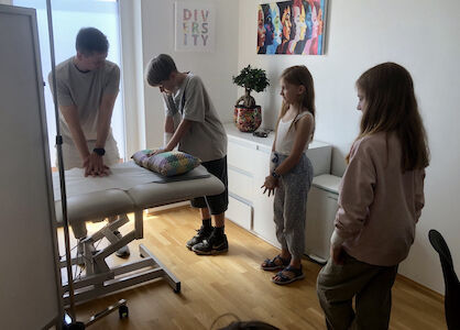 Drei Kinder schauen zu, wie ein Erwachsener auf einem Untersuchungstisch in einer Klinik eine Vorführung macht.