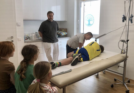 Ein Arzt untersucht das Bein eines Jungen, während andere in einem medizinischen Behandlungszimmer zuschauen.