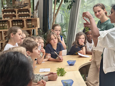Eine Gruppe von Kindern hört zwei Frauen zu, die an einem Tisch mit Pflanzen unterrichten.