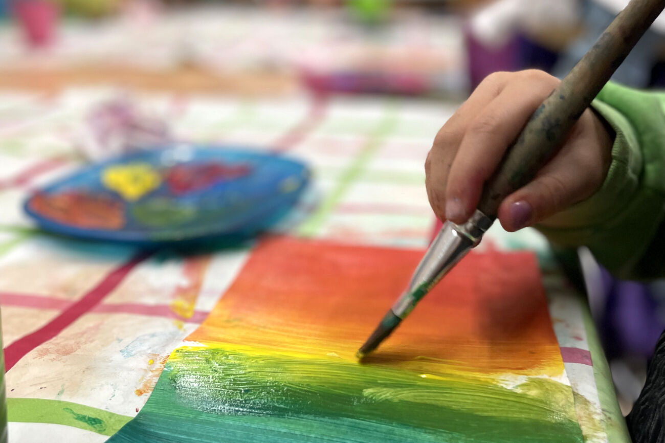 Zu sehen ist eine Kinderhand, die mit einem Borstenpinsel eine wunderschöne Farbabstufung malt. Im Hintergrund ist ein Farbmischteller zu sehen. Gearbeitet wird auf einer Plastikunterlage.