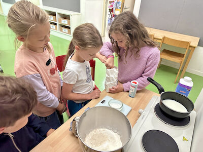 4 Kinder stehen vor einem Tisch und richten die Zutaten für die Kipferl her. Zwei Mädchen bemühen sich gerade den Kristallzucker genau abzuwiegen. Vor ihnen steht auch schon die erwärmte Milch im Topf auf der Herdplatte.