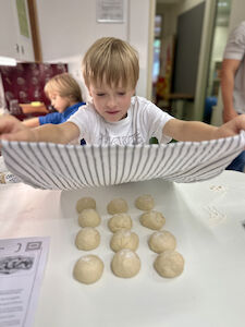 Ein Bub ist gerade dabei die fertig geformten Teigkugeln mit einem Geschirrtuch zuzudecken. Im Hintergrund wäscht sich ein anderer Bub gerade die Hände.