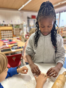 Ein Mädchen mit ganz vielen fein geflochtenen Zöpfen rollt die flach ausgewalkte Teigkugel zu einem Kipferl. Ein Bub unterstützt sie dabei.