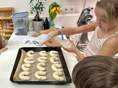 Ein Mädchen besprüht gerade die aufgegangenen Kipferl am Backblech mit einem transparenten Spritzfläschchen, bevor sie in den Backofen geschoben werden.