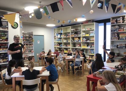 In einem Raum von Ludovico sitzen viele Kinder an Tischen und hören einem Erwachsenen zu, der etwas erklärt.