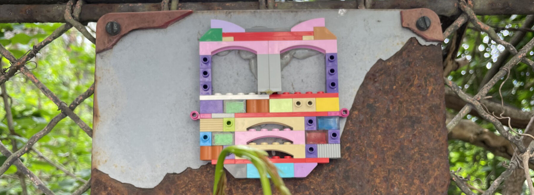 Auf einem Maschendrahtzaun hängt eine Tafel, wo aus Lego eine kleine Skulptur angebracht ist.