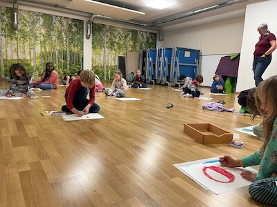 Mehrere Kinder malen am Parkettboden im Bewegungsraum ihren "perfekten Apfel". Im Hintergrund ist die Religionslehrerin Frau Hofer zu sehen.