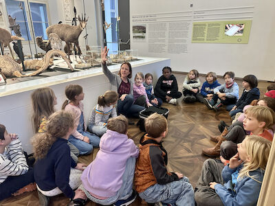 Die Führerin erklärt den Kindern etwas über Tarnung und Täuschung im Tierreich. Die Kinder sitzen im Kreis am Boden und hören aufmerksam zu.
