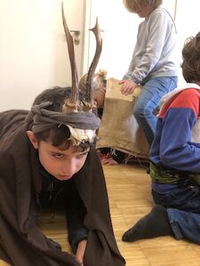 Im Vordergrund ist ein auf allen Vieren mit einem Geweih am Kopf zu sehen. Dahinter reitet ein Kind auf einem anderen, dessen Rücken mit einer Satteldecke bedeckt ist.