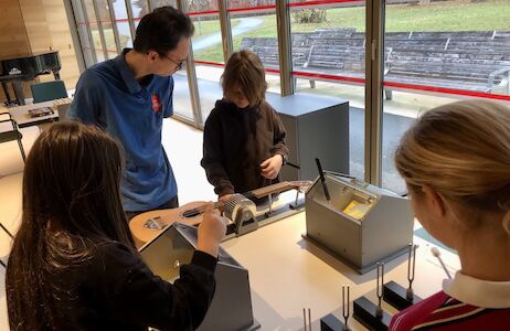 Ein Mitarbeiter vom Museum unterstützt die Kinder bei einer Musikstation, wo es um eine spannend adaptierte Gitarre geht.