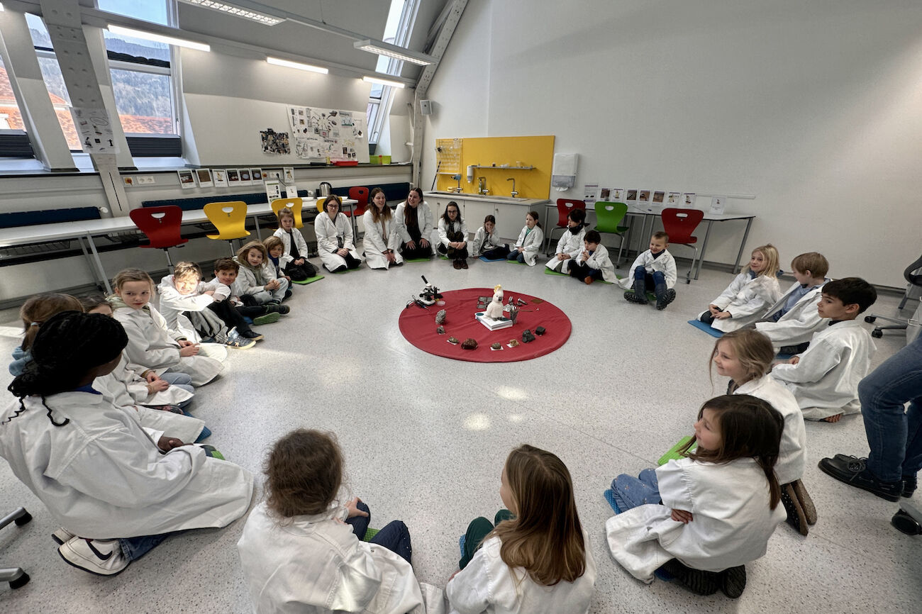 Die Kinder der Fuchs-Klasse sitzen mit ihren weißen Arbeitsmänteln im Kreis und hören den Ausführungen der Workshopleiterinnen zu.