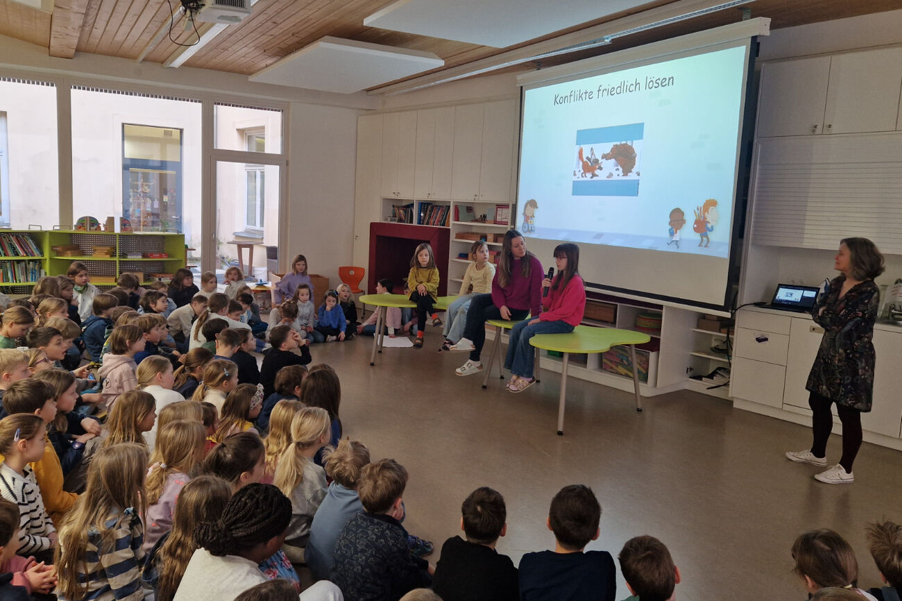 Ein Teil der Delfin-Klasse bei der Podiumsdiskussion im Rahmen der Schulversammlung. Drei Kinder sitzen am Tisch mit einer Lehrerin, im Hintergrund ist ein Bild auf der Leinwand zu sehen.