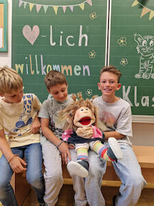 Drei Buben sitzen mit einer großen Stoffpuppe vor der Kreidetafel, worauf "Herzlich Willkommen" steht.
