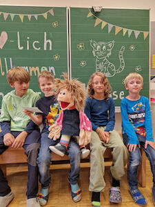 Vier Buben sitzen mit einer großen Stoffpuppe vor der Kreidetafel, worauf "Herzlich Willkommen" steht.