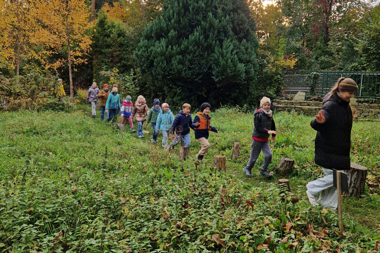 Im Gänsemarsch wird eine Wiese von Kindern lächelnd durchschritten. Als Erste geht eine erwachsene Person.