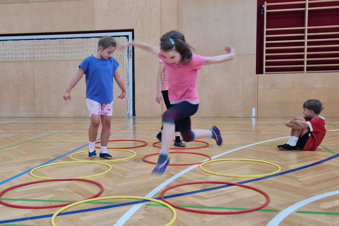 Ein Mädchen springt gerade von einem am Boden liegenden Reifen in einen anderen. Im Hintergrund sind weitere Kinder zu sehen.