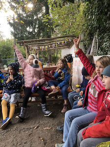 Die Kinder sitzen im Grünen im Kreis und zeigen vereinzelt auf.