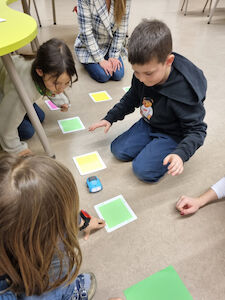Eine kleine Kindergruppe arbeitet mit Farbkärtchen und einem selbstfahrenden Auto am Boden.