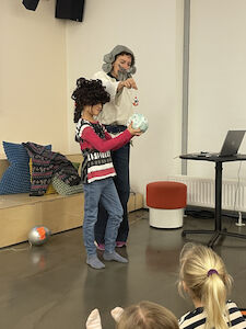 Eine verkleidete Erwachsene spielt mit einem Kind, das eine Perücke trägt die Geschichte von Münchhausen nach.