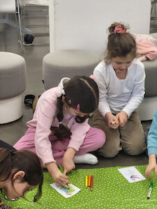 Mehrere Mädchen malen am Boden auf einer grünen Unterlage kleine Bilder mit Buntstiften aus.