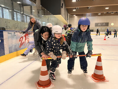 Eine Kindergruppe mit einer Betreuerin rutscht vergnügt auf Hütchen und einer Querstange gekonnt über die Eisfläche.