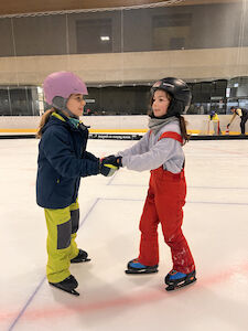 In Doppelhandhaltung bewegen sich zwei Kinder vorsichtig auf der Eisfläche.