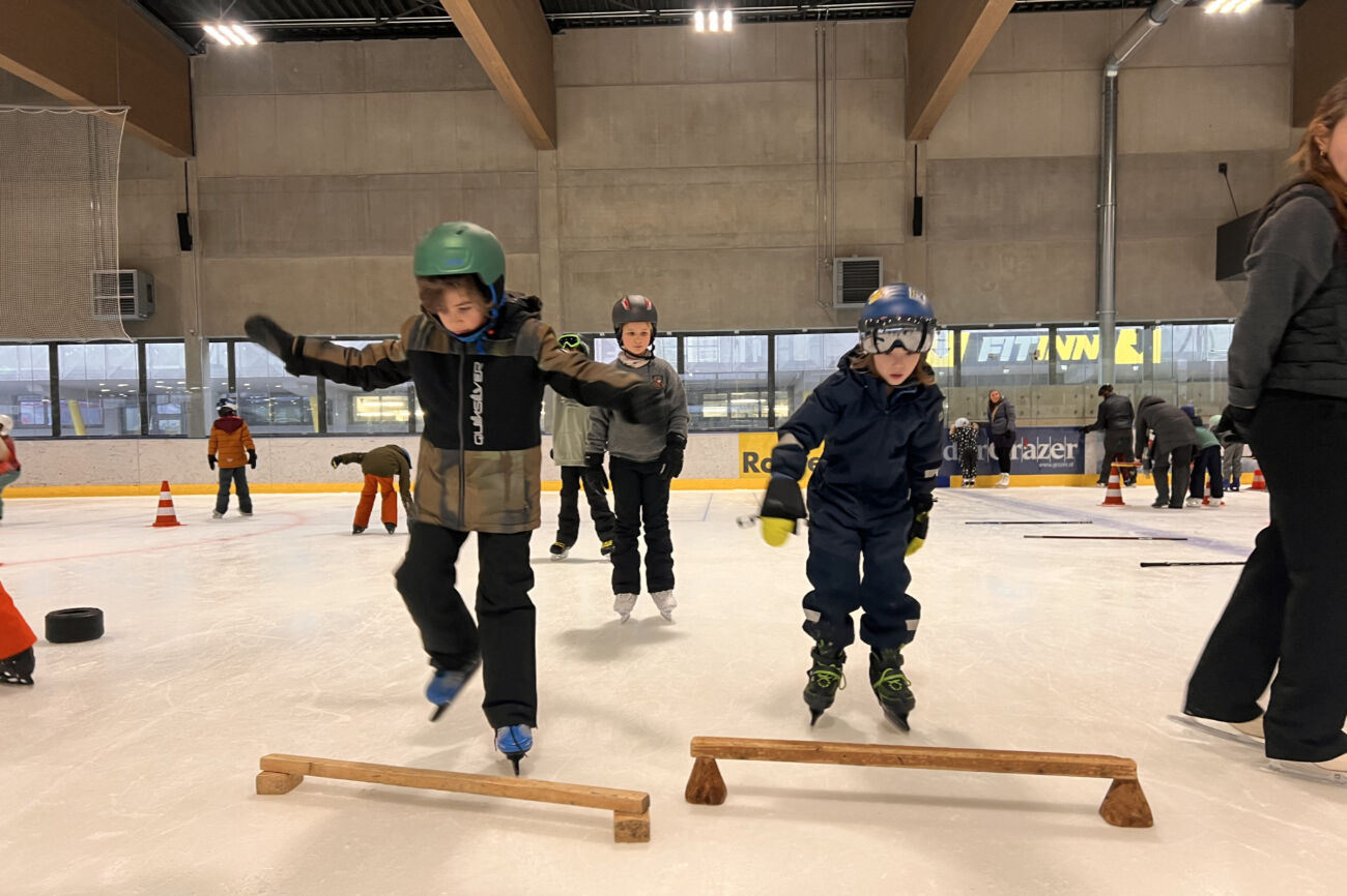 Zwei Buben probieren mit den Eislaufschuhen über ein Holzhindernis am Eis zu springen.