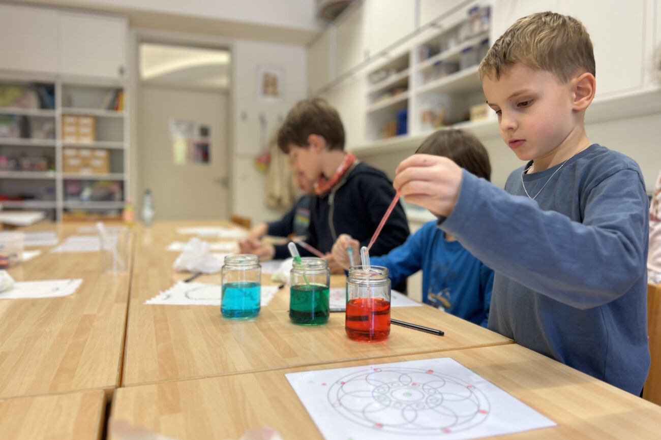 Ein Bub arbeitet mit einer Pipette und drei mit farbigem Wasser gefüllten Gläsern, um ein Muster auf ein laminiertes Blatt zu tropfen.