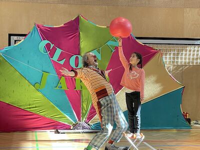 Clown Jako schaut mit ausgestreckten Armen begeistert einem Mädchen zu, das einen roten Ball auf einem Fingerangedreht balanciert.