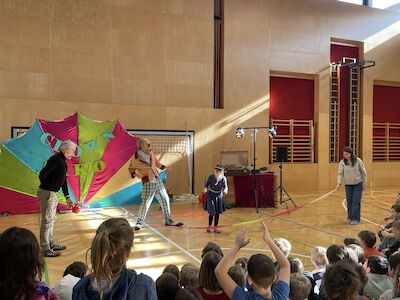 Clown Jako und zwei Lehrpersonen sind mit einem Schüler auf der Bühne im Turnsaal zu sehen, wie sie eine Riesenspringschnur drehen.