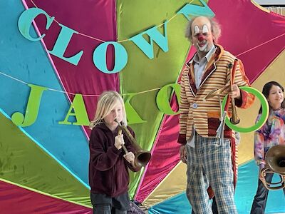 Ein Bub probiert sich mit einer Trompete und Clown Jako hält währenddessen mehrere Wurfringe in der Hand.