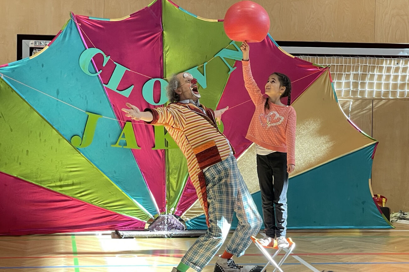 Clown Jako schaut mit ausgestreckten Armen begeistert einem Mädchen zu, das einen roten Ball auf einem Fingerangedreht balanciert.