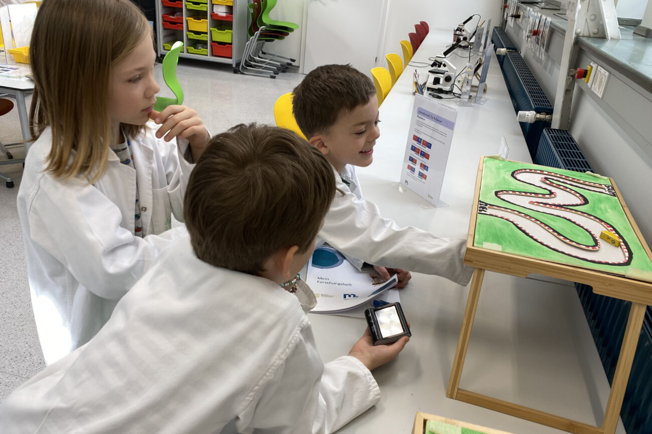 Drei Buben testen eine Rennautobahn der anderen Art aus, wo man von unten einen Magneten bewegt, um auf der Platte oben das Auto zu bewegen.