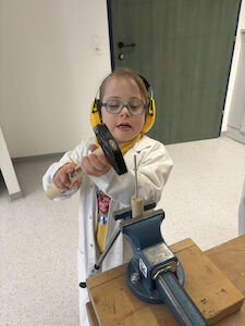 Ein Mädchen hämmert einen Nagel in ein Rundholz, das in einem Schraubstock eingespannt ist.