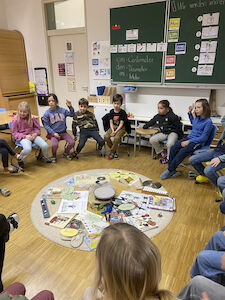 Die Kinder sitzen im Sesselkreis um den Teppich in der Klasse, worauf Vieles über Musik aufgelegt ist.