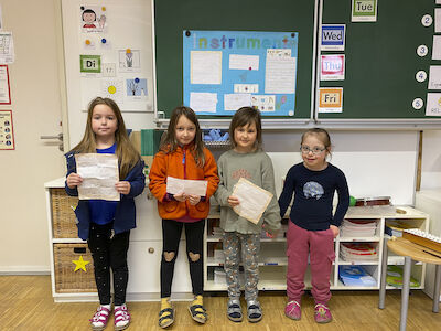 Eine Mädchengruppe präsentiert ein Referat über Instrumente vor der Tafel. Hinter ihnen haben sie ihr Plakat aufgehängt.