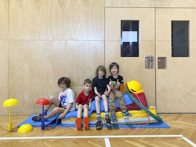 Einige Buben sitzen auf mehreren Schaumstoffteilen, die auf einer blauen Matte liegen und lachen in die Kamera.