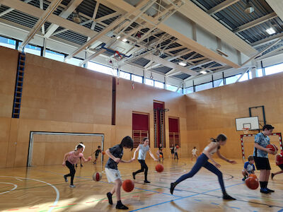 Bei einem Basketball-Spiel sind viele Kinder aktiv im Turnsaal unterwegs.