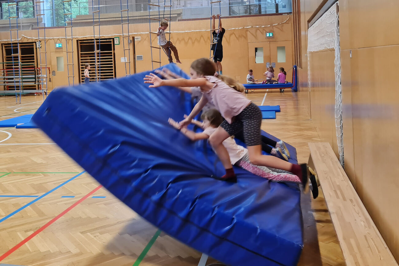 Mehrere Kinder fallen gemeinsam mit der dicken, aufgestellten Matte im Turnsaal auf den Boden.